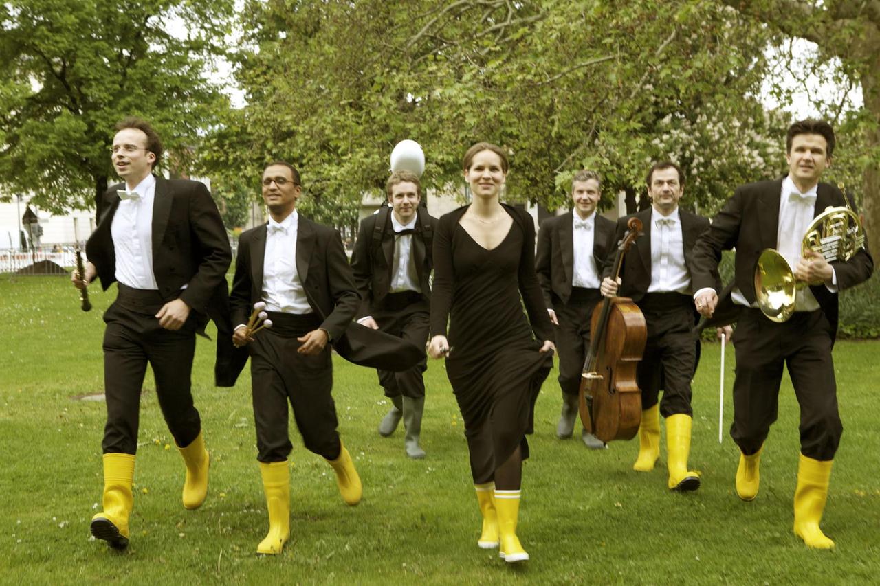 Sieben Musiker in Anzügen und mit gelben Gummistiefeln laufen über eine Wiese in Richtung Kamera