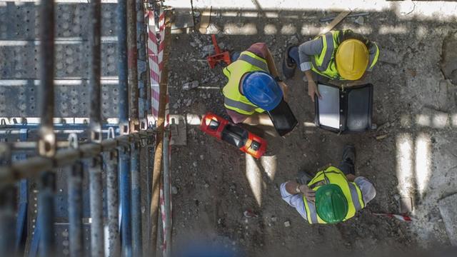 Blick von oben auf drei Bauarbeiter mit Schutzhelmen, die auf einer Baustelle zusammenstehen
