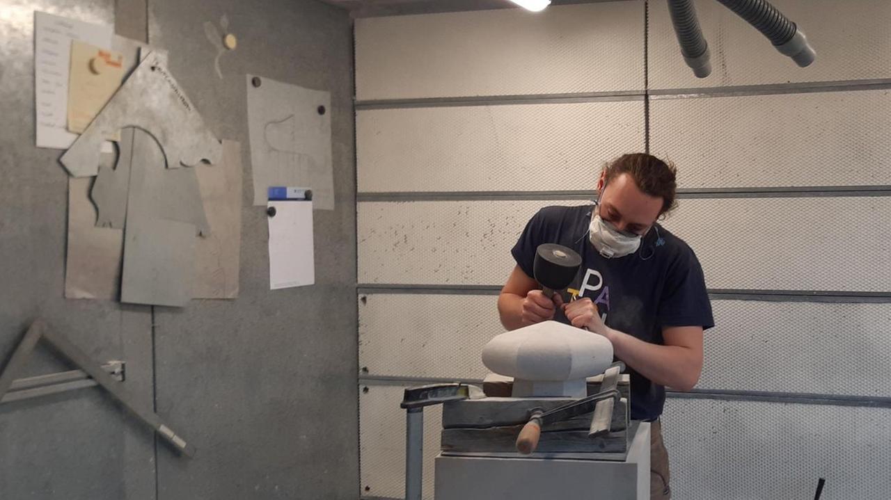 Charles meißelt per Hand an einem Stein- ohne technische Hilfsmittel. Er trägt die Haare nach hinten gebunden, eine FFP2 Maske und blickt konzentriert auf den Stein, den er bearbeitet. Charles meißelt per Hand an einem Stein- ohne technische Hilfsmittel. Er trägt die Haare nach hinten gebunden, eine FFP2 Maske und blickt konzentriert auf den Stein, den er bearbeitet.