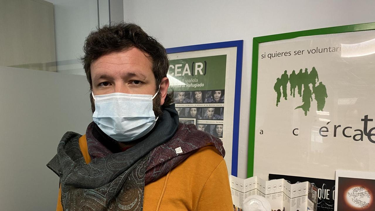 Ein junger Mann mit dunklem Haar, gelbem Pulli und Mundschutz sowie einem Halstuch steht in seinem Büro. Ein junger Mann mit dunklem Haar, gelbem Pulli und Mundschutz sowie einem Halstuch steht in seinem Büro.