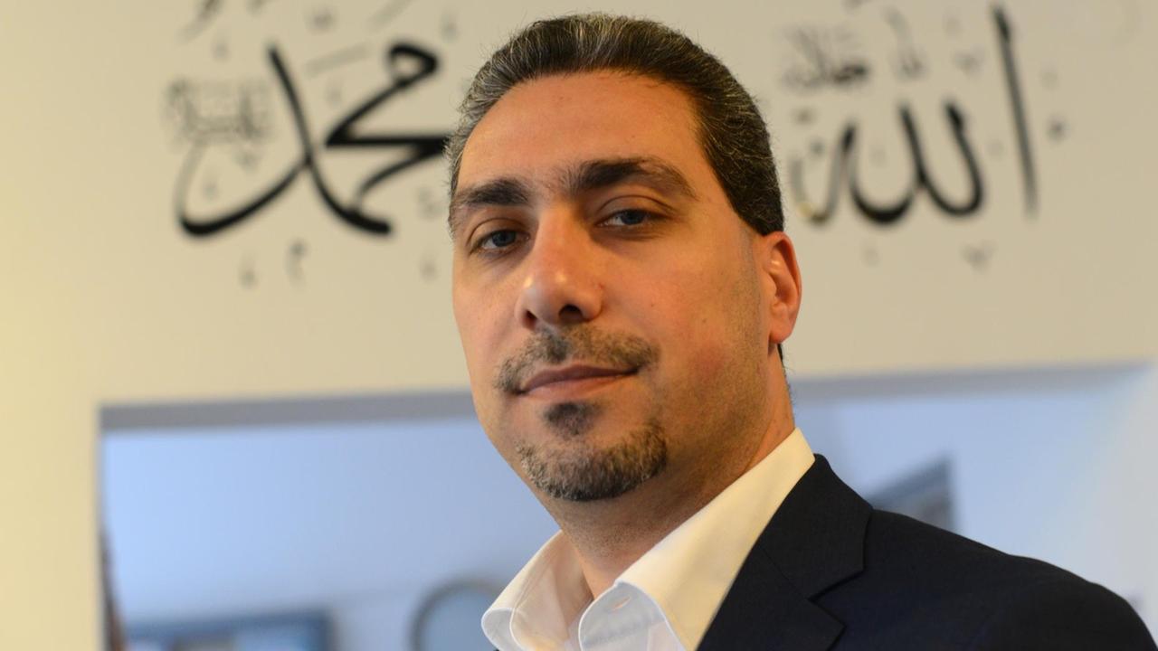 Sadiqu Al-Mousllie im schwarzen Sakko und weißem Hemd vor einem Türeingang mit arabischen Schriftzügen. Er blickt in die Kamera. Sadiqu Al-Mousllie im schwarzen Sakko und weißem Hemd vor einem Türeingang mit arabischen Schriftzügen. Er blickt in die Kamera.
