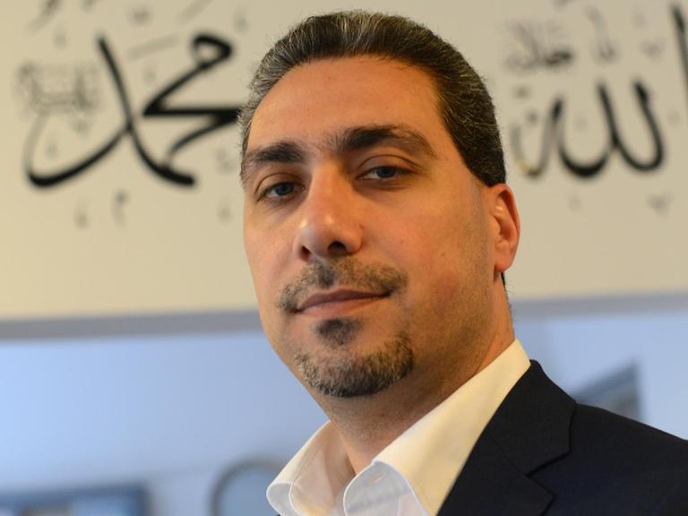 Sadiqu Al-Mousllie im schwarzen Sakko und weißem Hemd vor einem Türeingang mit arabischen Schriftzügen. Er blickt in die Kamera.