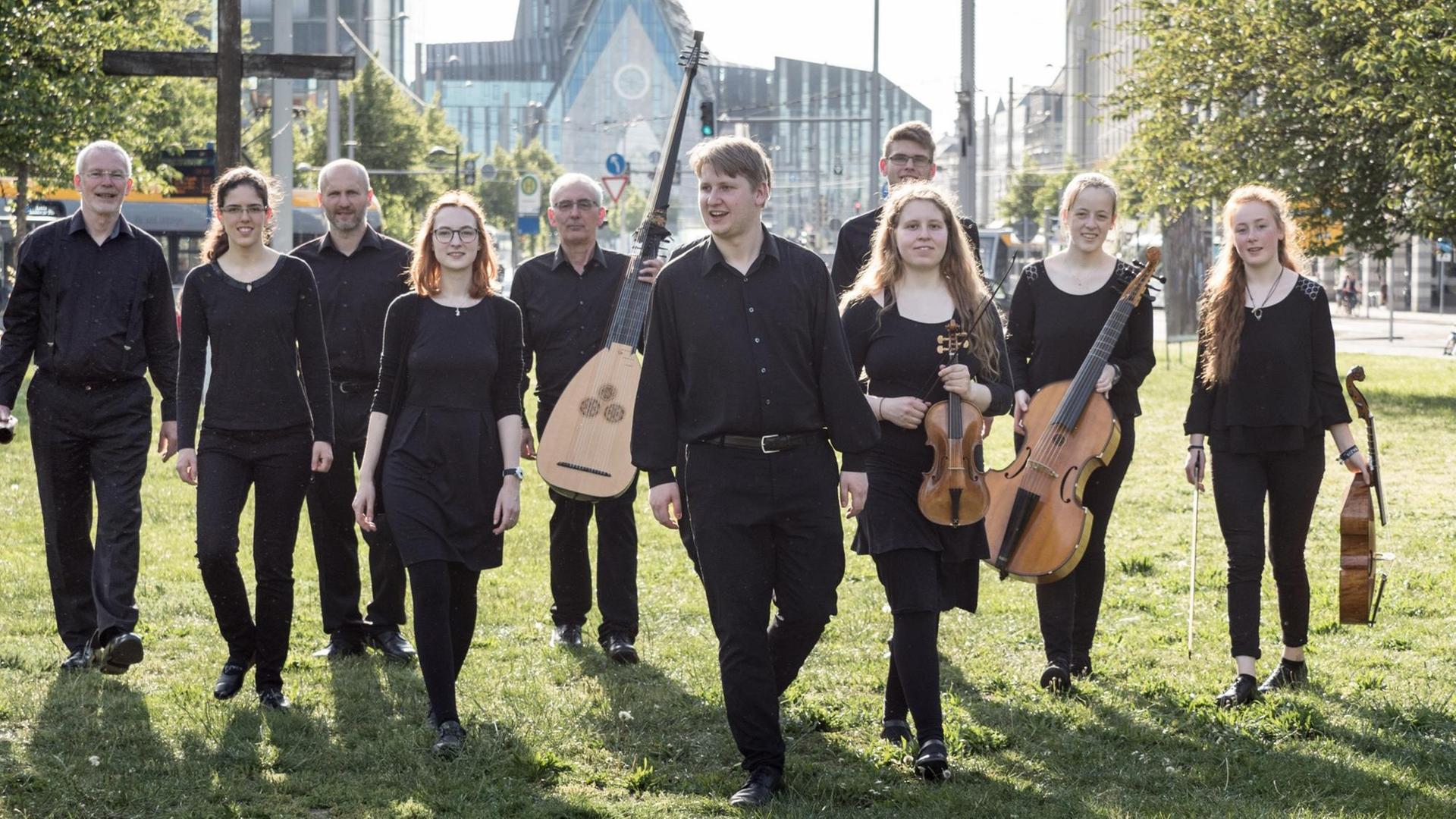 Das Ensemble in schwarzer, legerer Konzertkleidung befindet sich auf einer grünen Wiese in der Stadt, manche halten ihre Streich- und Zupf-Instrumente in der Hand, sie laufen auf den Fotografierenden zu. Abgebildet sind 10 Personen. Das Ensemble in schwarzer, legerer Konzertkleidung befindet sich auf einer grünen Wiese in der Stadt, manche halten ihre Streich- und Zupf-Instrumente in der Hand, sie laufen auf den Fotografierenden zu. Abgebildet sind 10 Personen.