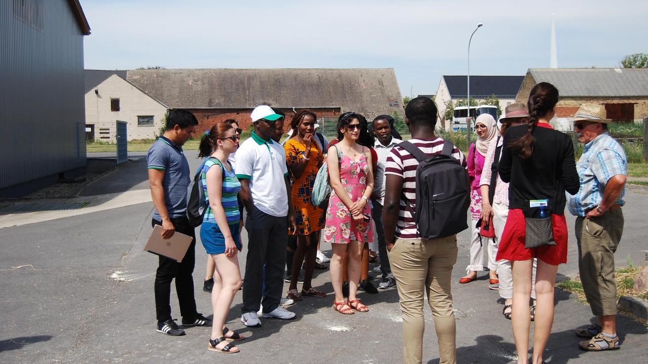 Eine Besuchergruppe in sommerlicher Kleidung steht in Feldheim auf der Straße und hört den Ausführungen von Kathleen Thomson zu. Eine Besuchergruppe in sommerlicher Kleidung steht in Feldheim auf der Straße und hört den Ausführungen von Kathleen Thomson zu.