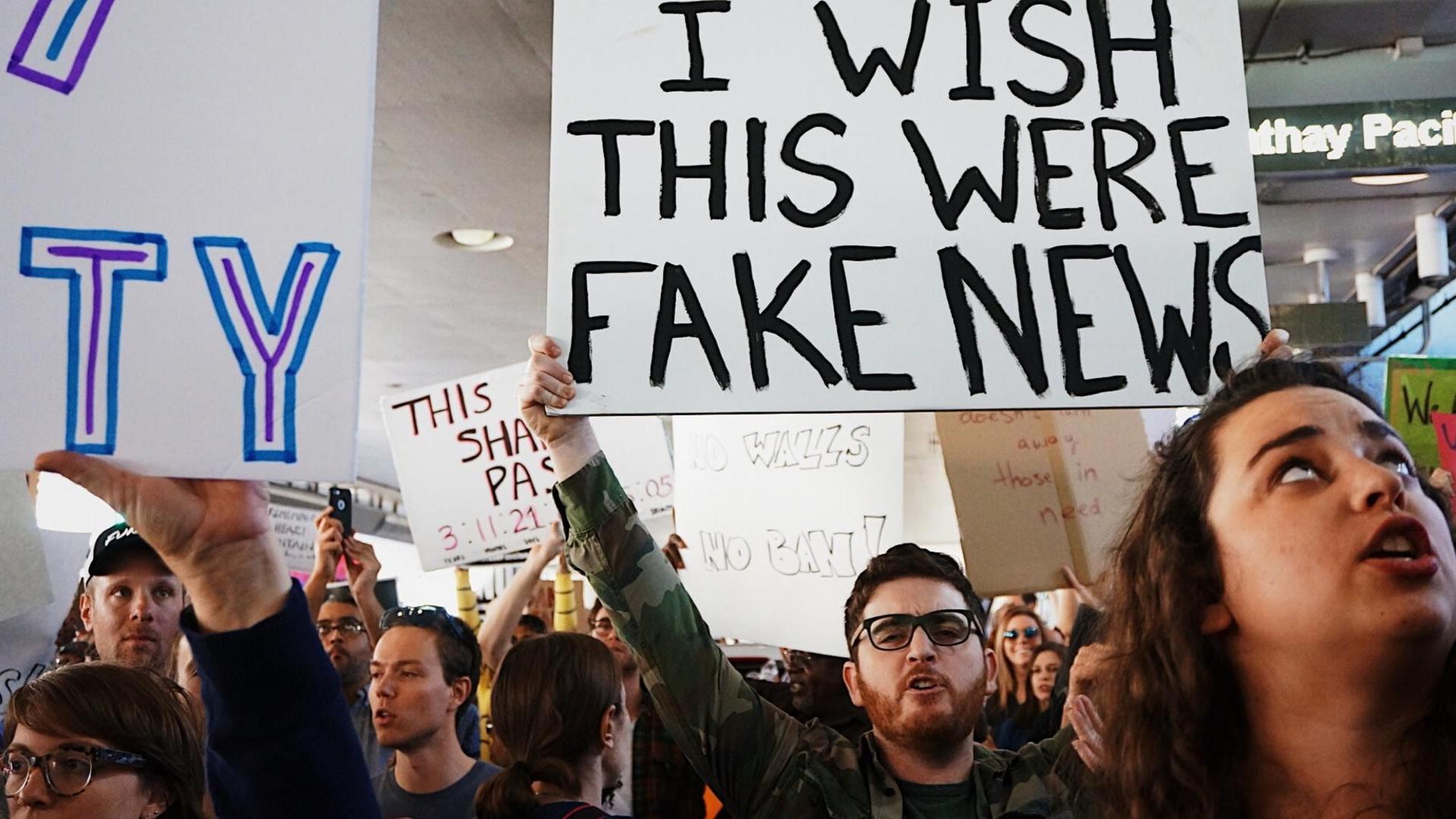 Menschen demonstrieren. Ein Mensch hält ein Schild in die Höhe auf dem auf Englisch steht: "Ich wünschte das wären Fake News."