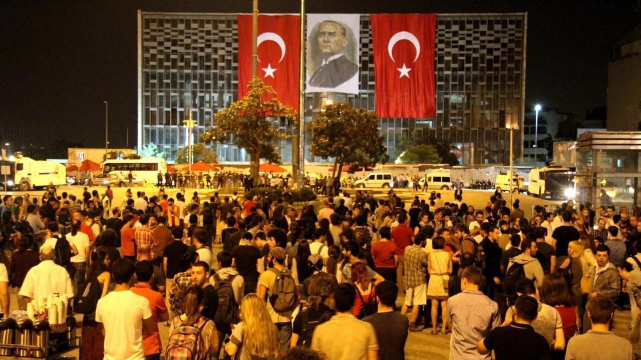 Auf dem nächtlichen Taksim Square in Istanbul stehen viele junge Menschen und schauen auf das Atatürk-Kulturzentrum, an dessen Fassade zwei türkische Flaggen und ein Porträt Atatürks angebracht sind.