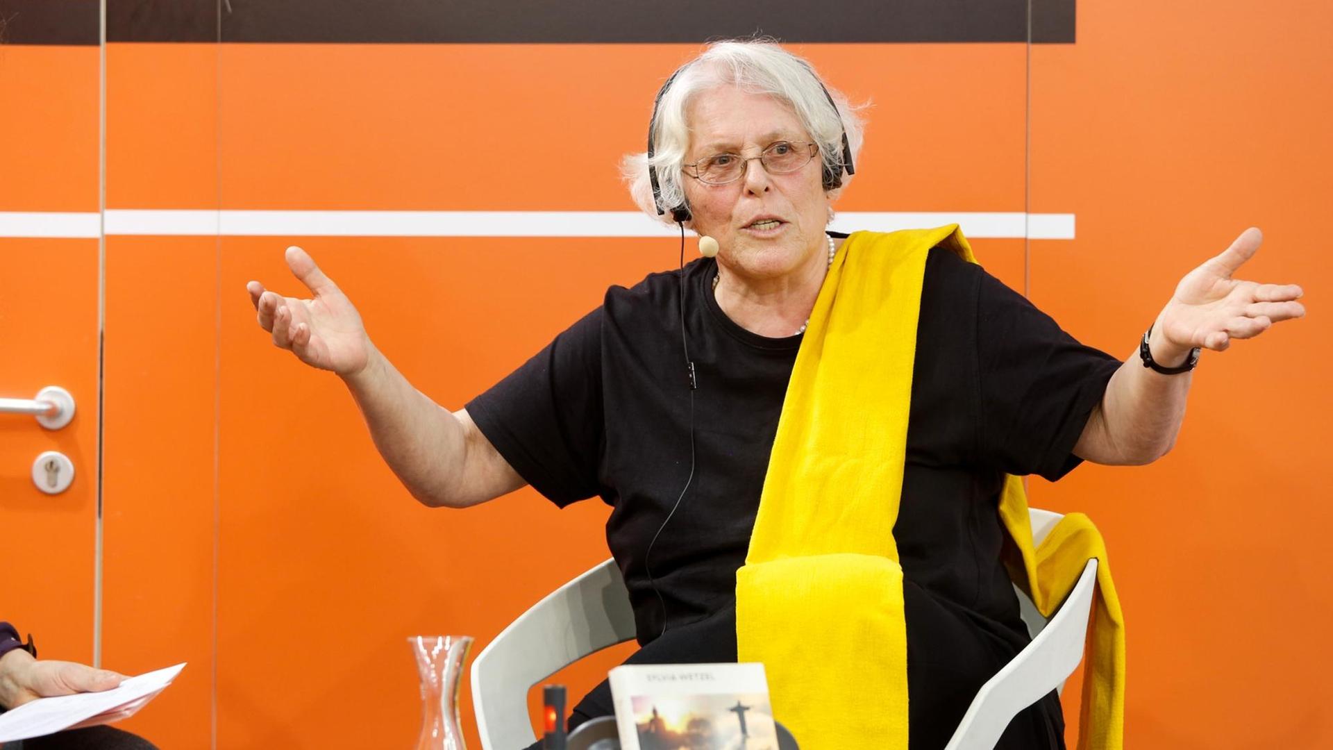 Syvia Wetzel auf der Bühne von Deutschlandfunk Kultur, mit Mikrofon und gestikulierend. Syvia Wetzel auf der Bühne von Deutschlandfunk Kultur, mit Mikrofon und gestikulierend.