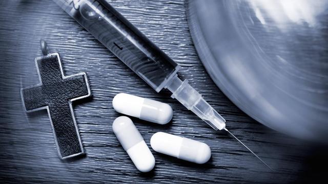 Wasserglas, Kreuz, Tabletten und Spritze auf einem schwarz-weißen Foto.