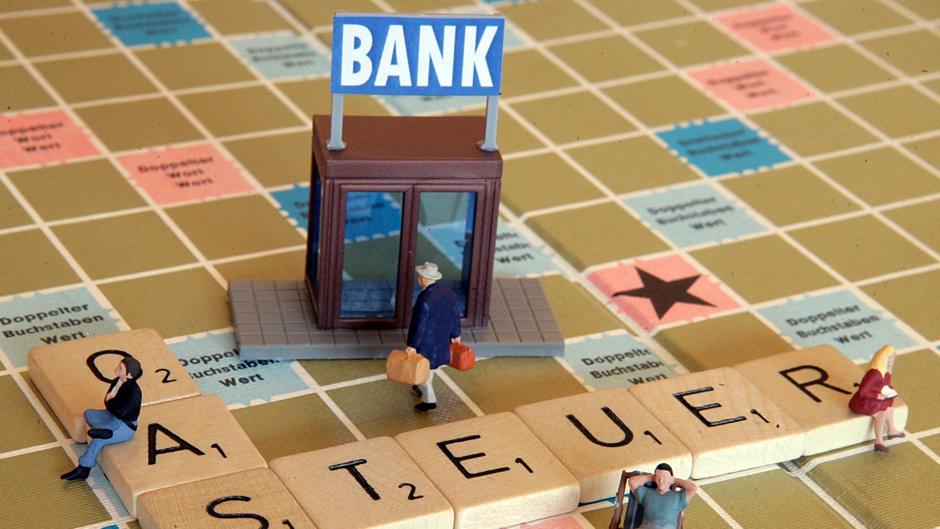 Eine Miniaturfigur trägt zwei Geldkoffer auf einem Scrabble-Spiel in eine Bank (Illustration), auf dem das Wort Steueroase liegt.