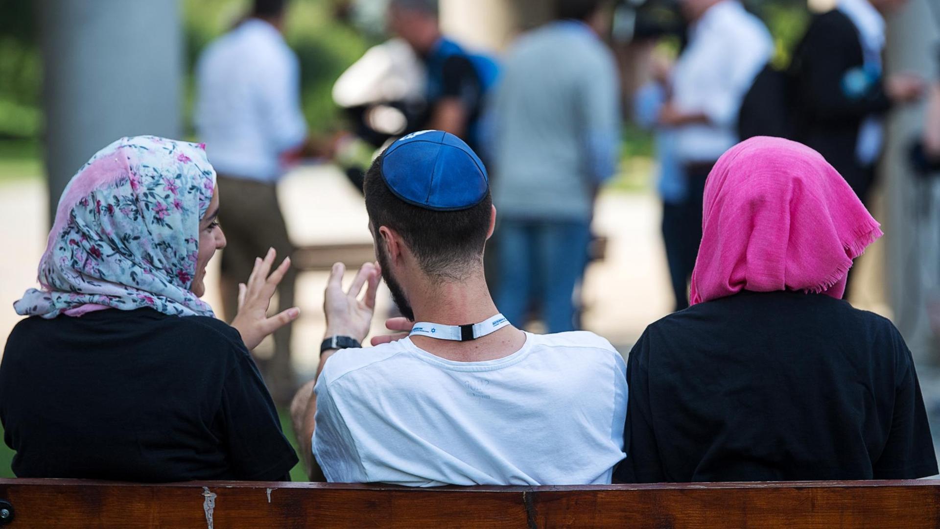 Zwei junge Muslima mit Kopftuch unterhalten sich auf einer Bank sitzend mit einem jungen jüdischen Mann mit Kippa. Zwei junge Muslima mit Kopftuch unterhalten sich auf einer Bank sitzend mit einem jungen jüdischen Mann mit Kippa.