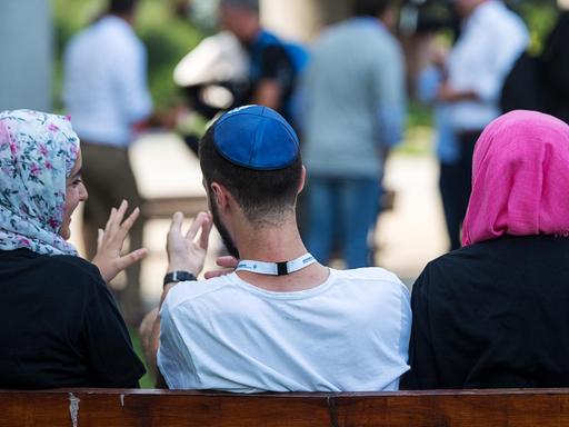 Zwei junge Muslima mit Kopftuch unterhalten sich auf einer Bank sitzend mit einem jungen jüdischen Mann mit Kippa.