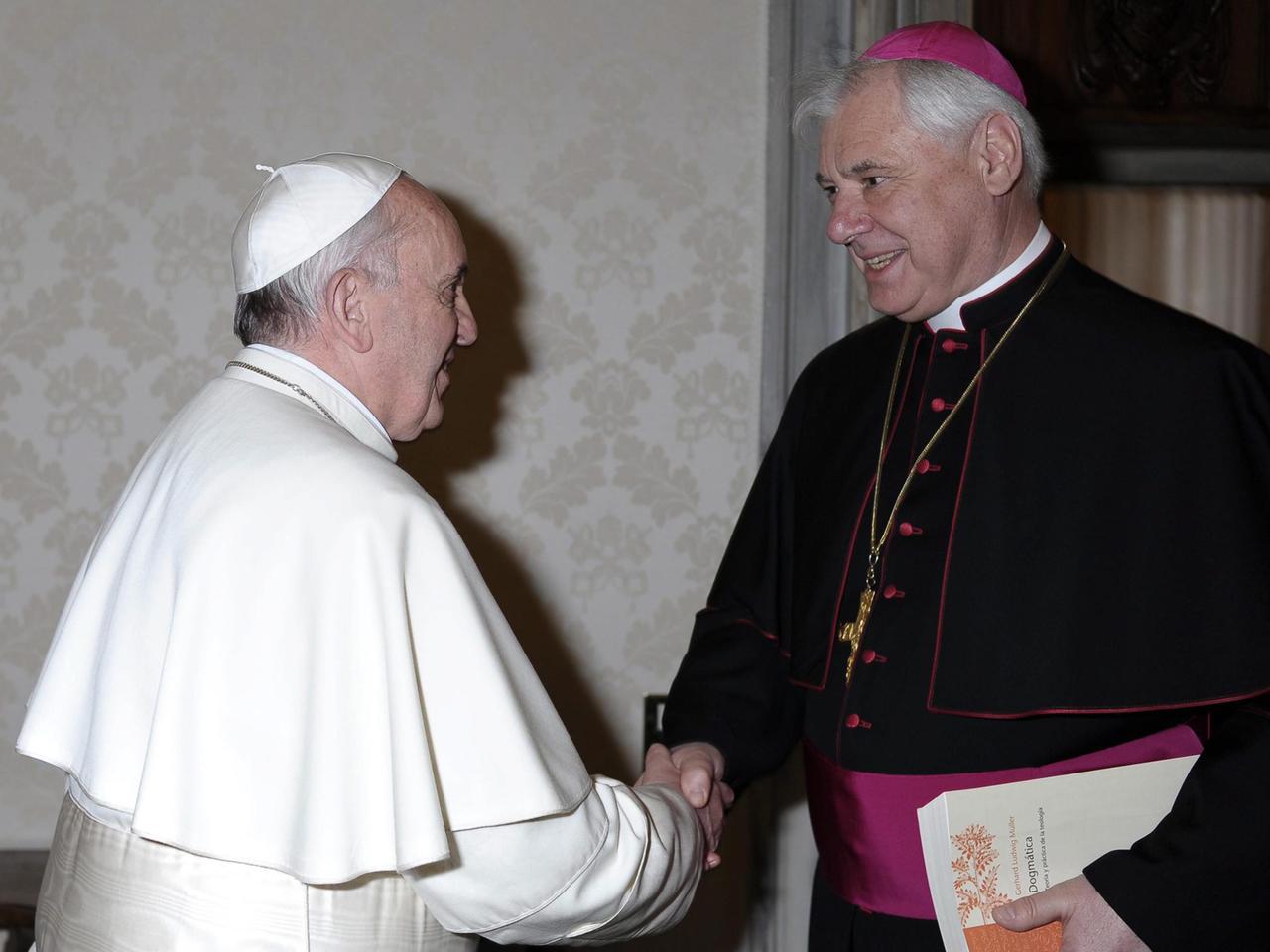 Papst Franziskus, ganz in weiß gekleidet, schüttelt dem Präfekten der Glaubenskongregation, Gerhard Ludwig Müller, die Hand.