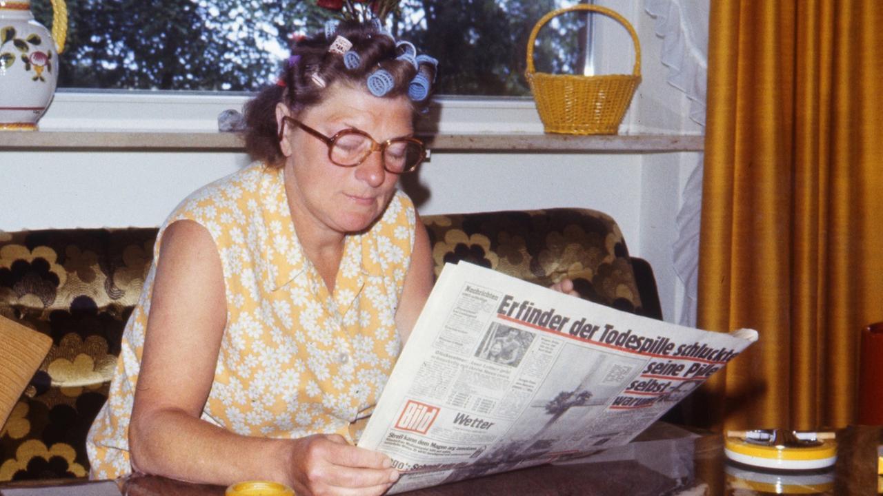Eine Frau mit Lockenwicklern im Haar und in Kittelschürze sitzt Ende der 70er-Jahre auf einer Couch und liest eine Zeitung. Eine Frau mit Lockenwicklern im Haar und in Kittelschürze sitzt Ende der 70er-Jahre auf einer Couch und liest eine Zeitung.