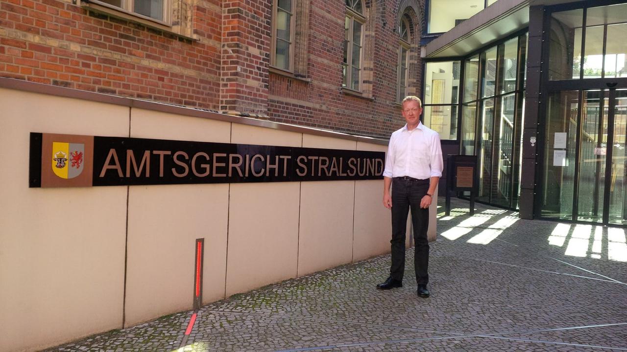 Axel Peters, Mann in schwarzer Hose und weißem Hemd, steht vor einem Gebäude mit dem Schild "Amtsgericht Stralsund":