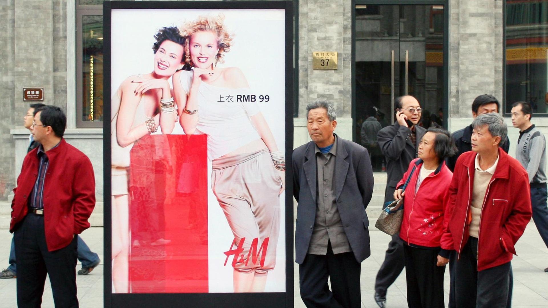 Chinesen und Chinesinnen stehen auf dem Gehweg neben einer Werbung des Modeherstellers H&M.