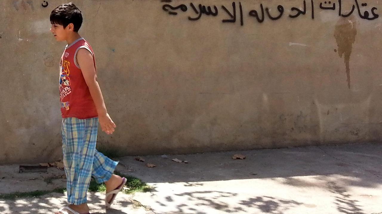 Der Junge trägt ein rotes Shirt und läuft auf einem Bürgersteig an der Hauswand vorbei. Das Graffito ist mit schwarzer Farbe gesprüht.
Der Junge trägt ein rotes Shirt und läuft auf einem Bürgersteig an der Hauswand vorbei. Das Graffito ist mit schwarzer Farbe gesprüht.