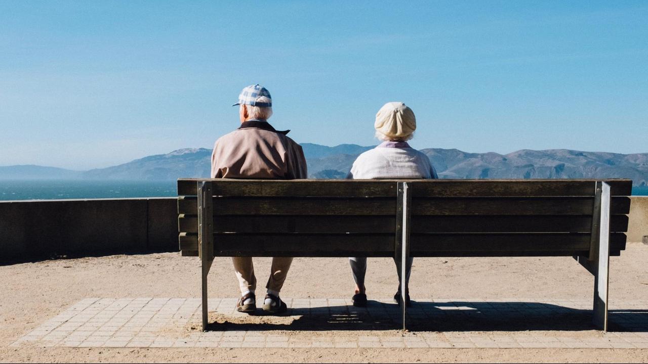 Zwei ältere Leute, von hinten gesehen, sitzen nebeneinander auf einer Bank und schauen auf's Meer.