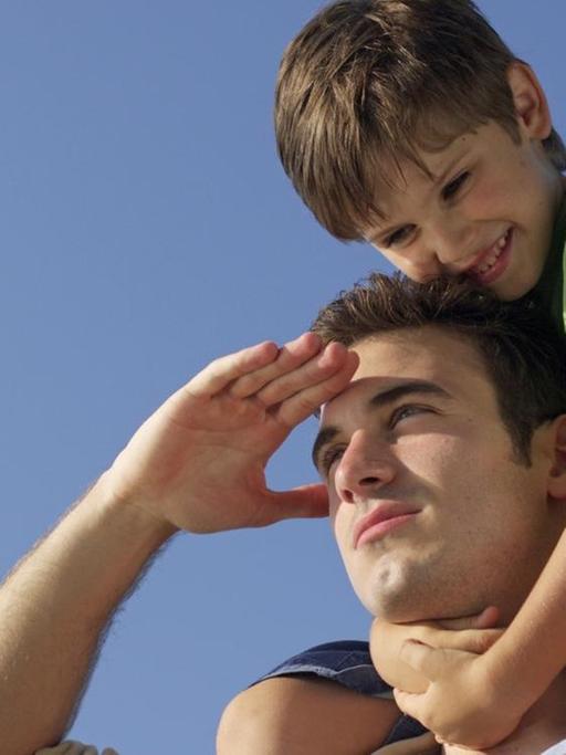 Ein Junge sitzt auf den Schultern seines Vaters, der seine Hand an der Stirn hat und in die Ferne blickt.