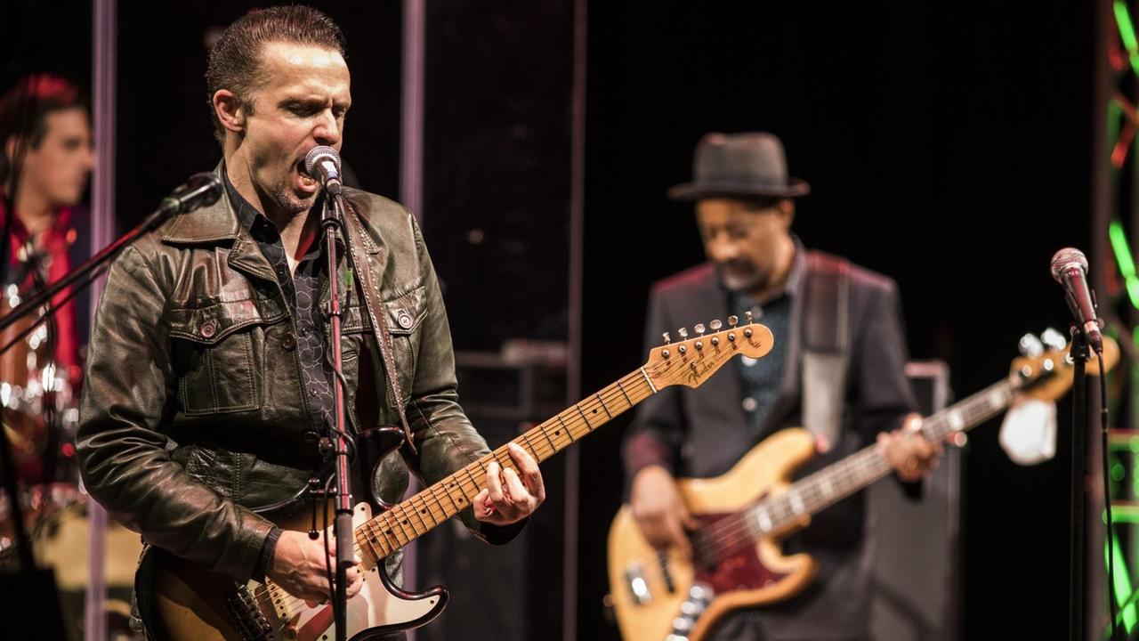 Drei Männer stehen auf einer Bühne: Links hinten ein Schlagzeuger, davor ein Gitarrist, der in ein Mikrofon singt, rechts ein Bassist mit Hut auf dem Kopf. Drei Männer stehen auf einer Bühne: Links hinten ein Schlagzeuger, davor ein Gitarrist, der in ein Mikrofon singt, rechts ein Bassist mit Hut auf dem Kopf.