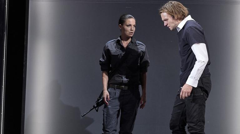 Eine Frau in dunkler Uniform und mit einem Maschinengewehr spricht mit einem Mann in Zivil. 