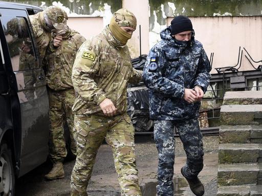 Zu sehen ist ein ukrainischer Marinesoldat, der in Handschellen von einem Offizier des russischen Nachrichtendienstes FSB zu einem Gerichtssaal begleitet wird.