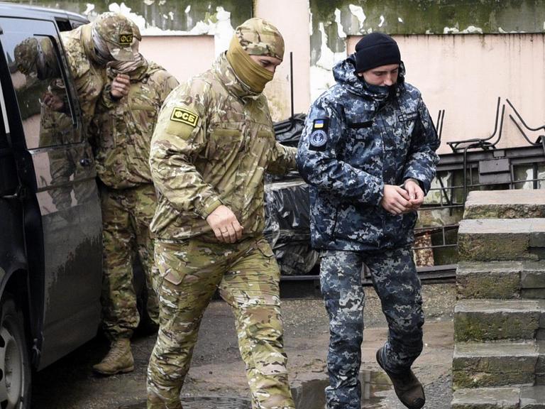 Zu sehen ist ein ukrainischer Marinesoldat, der in Handschellen von einem Offizier des russischen Nachrichtendienstes FSB zu einem Gerichtssaal begleitet wird.