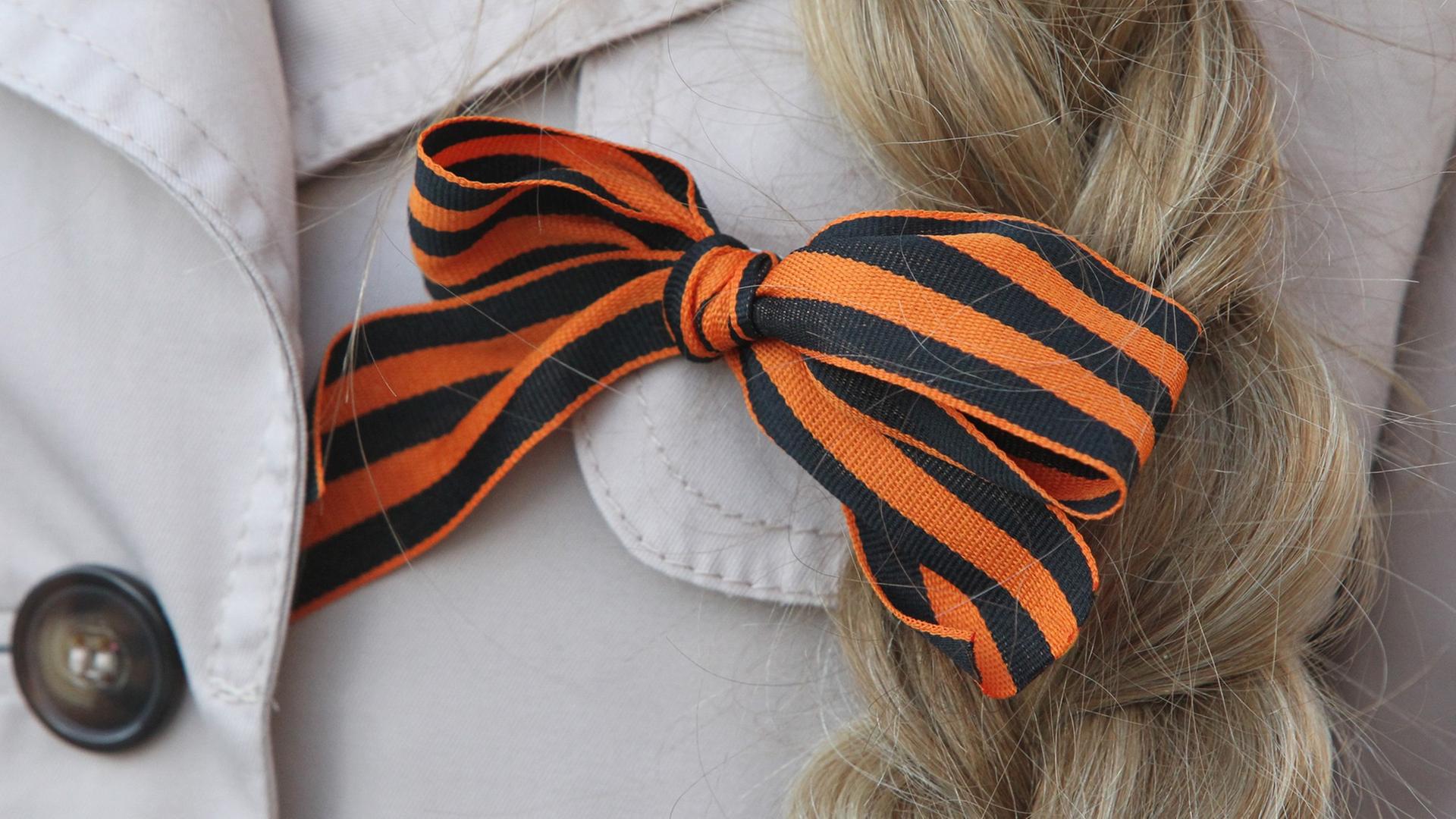 Großaufnahme des orange-schwarzen Georgsbands, hier zur Schleife gebunden am Revers einer Frau mit blondem geflochtenen Zopf. Großaufnahme des orange-schwarzen Georgsbands, hier zur Schleife gebunden am Revers einer Frau mit blondem geflochtenen Zopf.