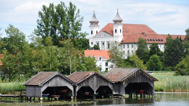 Zu sehen sind Teile des ehemaligen Klosters Schlehdorf am Kochelsee: Die große Pfarrkirche und ein Seitenflügel des Klosters, im Vordergrund sind Bootshäuser zu sehen.