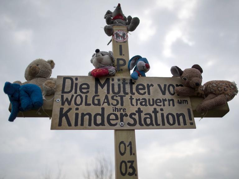 Ein Holzkreuz mit Plüschtieren und dem Schriftzug "Die Mütter von Wolgast trauern um ihre Kinderstadtion!" ist am 29.03.2017 am Ortsrand von Wolgast (Mecklenburg-Vorpommern) zu sehen.