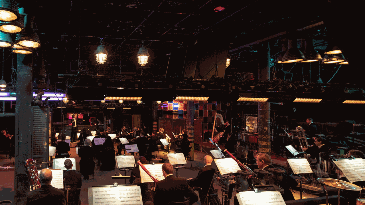 Das Orchester sitzt in einem Industrieraum und spielt nach Vorgabe seines Dirigenten.