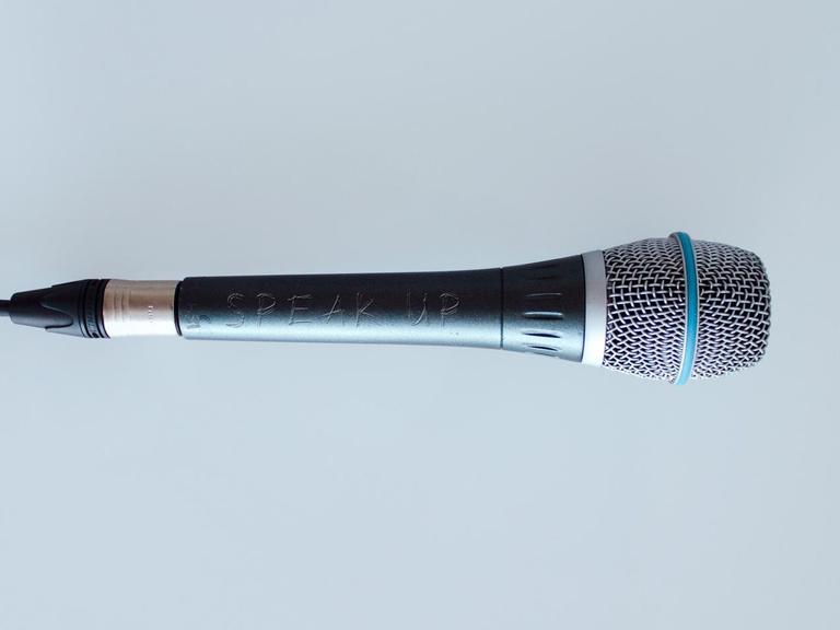 Ein Mikrofon vor grauem Hintergrund. Darauf eingeritz steht "Speak Up".