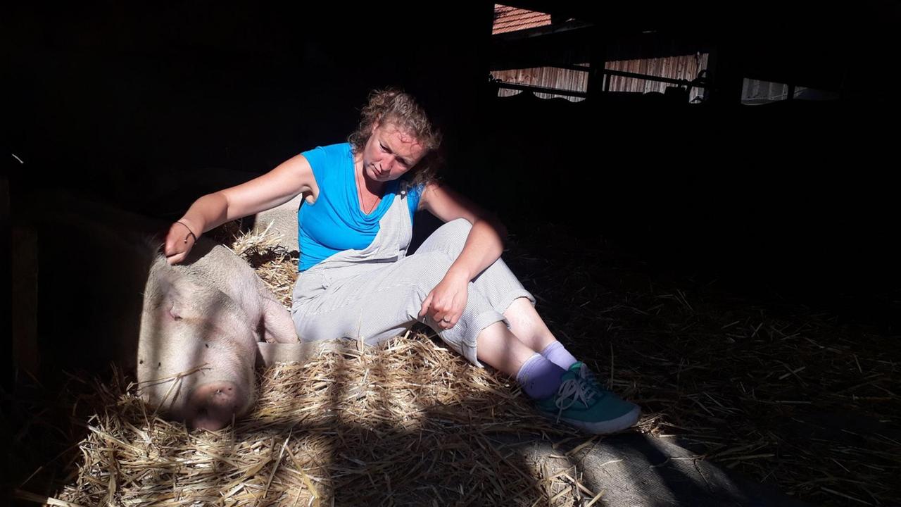 Eine blonde Frau sitzt in einem Stall auf Stroh. Neben ihr liegt ein Schwein.