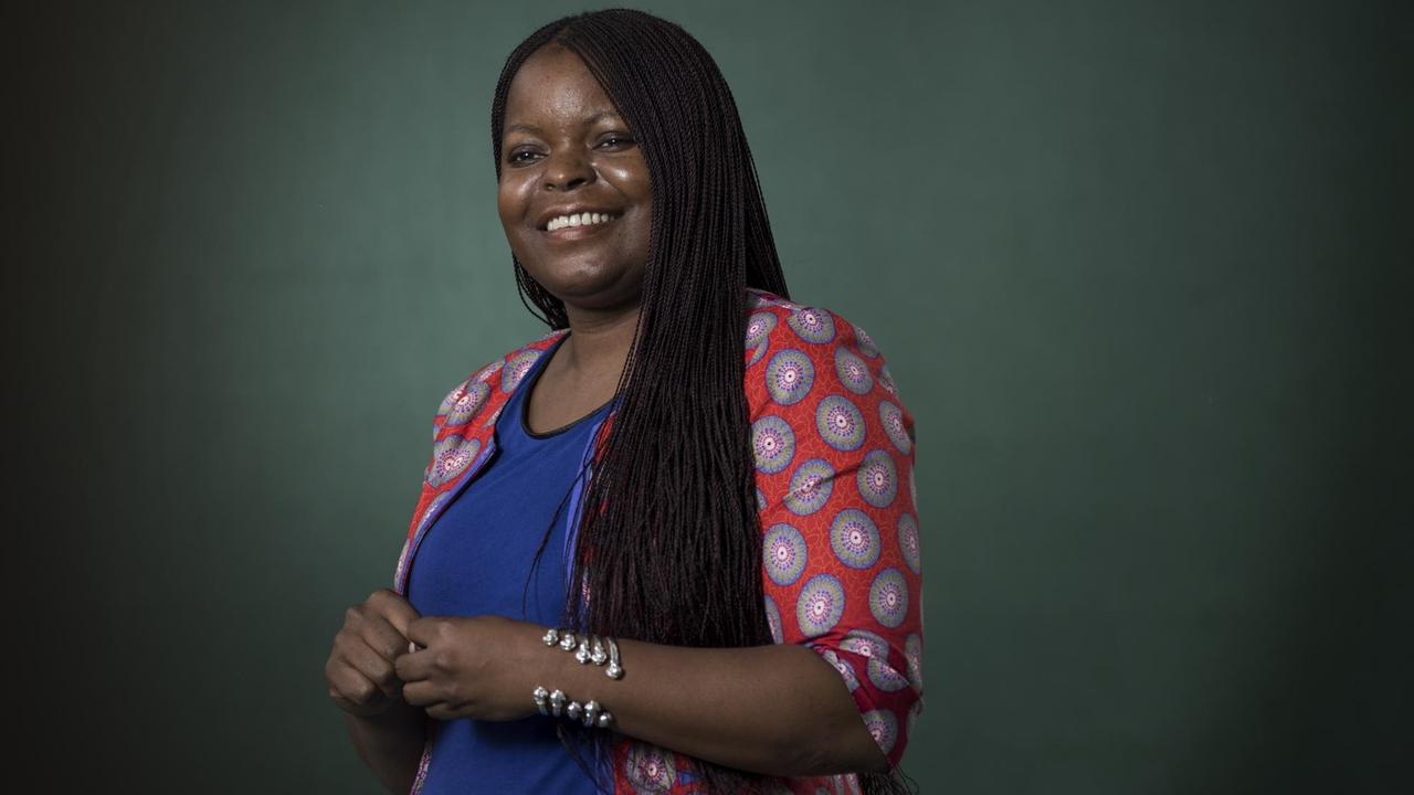 Petina Gappah steht vor dunkelgrünem Hintergrund, lächelt leicht und schaut links seitlich an der Kamera vorbei. Sie trägt ein dunkelblaues T-Shirt, einen rot-bunt gemusterten Blazer und einen silbernen Armreif um den linken Arm. Petina Gappah steht vor dunkelgrünem Hintergrund, lächelt leicht und schaut links seitlich an der Kamera vorbei. Sie trägt ein dunkelblaues T-Shirt, einen rot-bunt gemusterten Blazer und einen silbernen Armreif um den linken Arm.