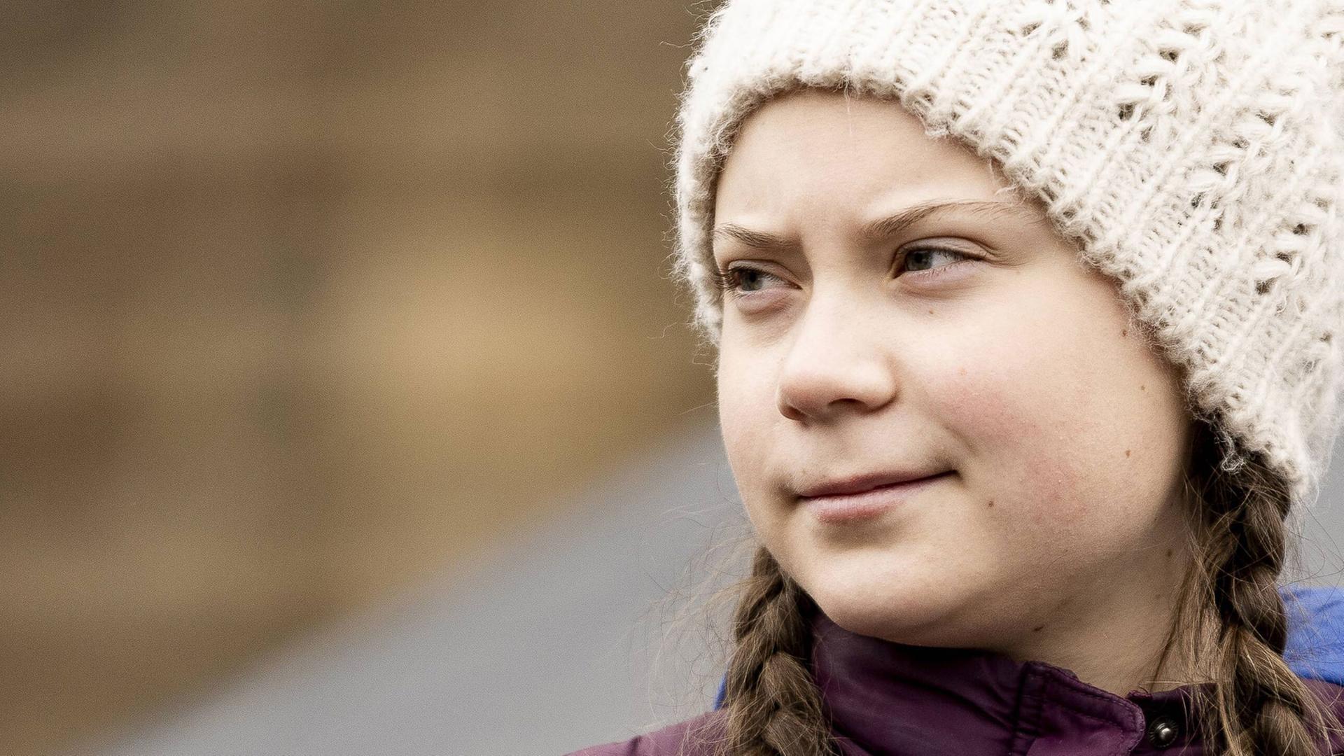 Greta Thunberg im Halbprofil, sie schaut mit ernstem Gescihtsausdruck zur Seite und trägt eine helle Mütze, aus der geflochtene Zöpfe herausschauen Greta Thunberg im Halbprofil, sie schaut mit ernstem Gescihtsausdruck zur Seite und trägt eine helle Mütze, aus der geflochtene Zöpfe herausschauen