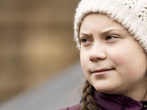 Greta Thunberg im Halbprofil, sie schaut mit ernstem Gescihtsausdruck zur Seite und trägt eine helle Mütze, aus der geflochtene Zöpfe herausschauen