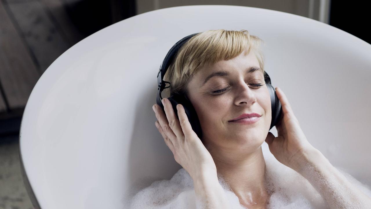 Ein blonde Frau liegt in einer weißen Badewanne mit Schaum, auf den Ohren trägt sie einen Kopfhörer.