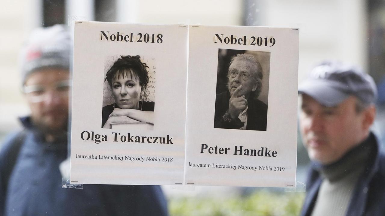 Zwei Blätter mit den Fotos der beiden Nobelpreisträger Olga Tokarczuk und Peter Handke hängen an der Glasscheibe einer Buchhandlung in Warschau. Durch die Glasscheibe sieht man zwei Passanten auf der Strasse.
