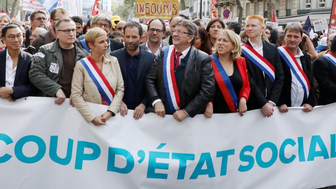 Jean-Luc Melenchon steht mit einer blau-weiß-roten Schärpe in einer Traube von Menschen, die gemeinsam einen weißen Banner mit der Aufschrift "Coup D'état Social" halten