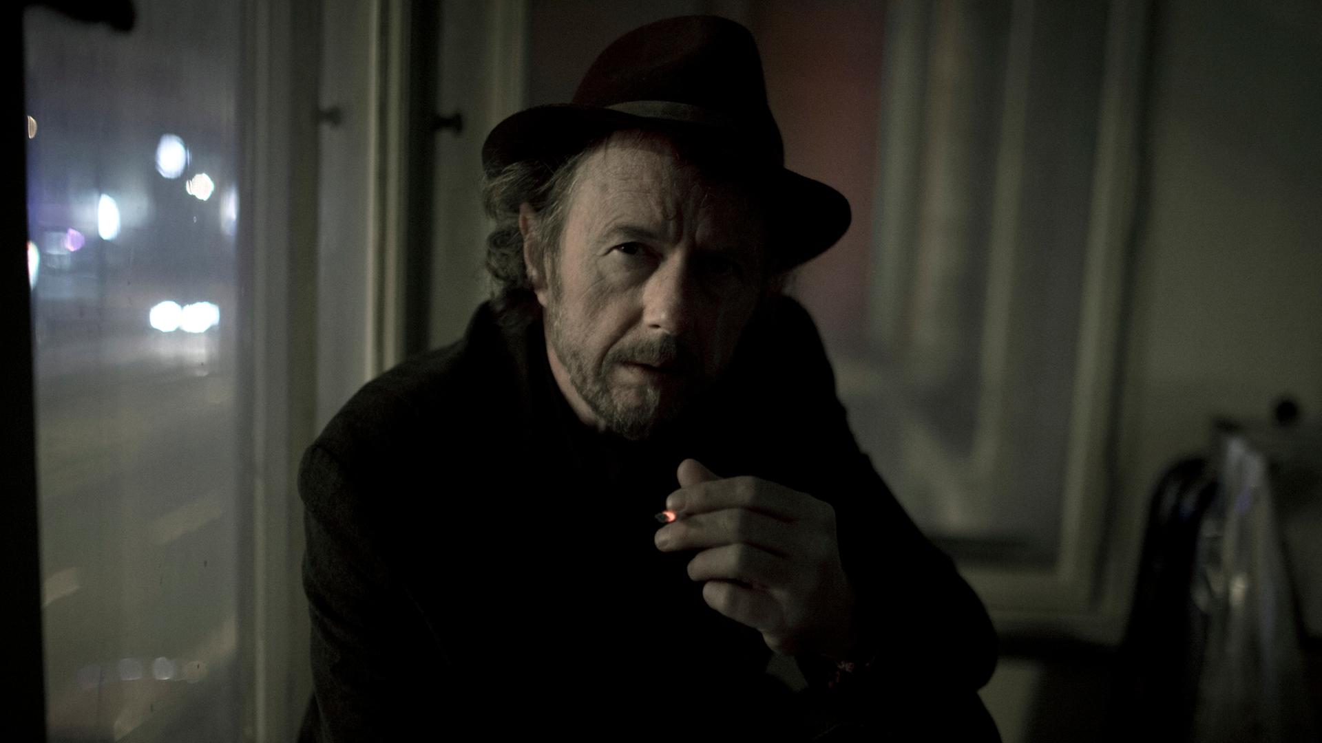Der Filmemacher Uli Schueppel hält eine Zigarette in den Händen.