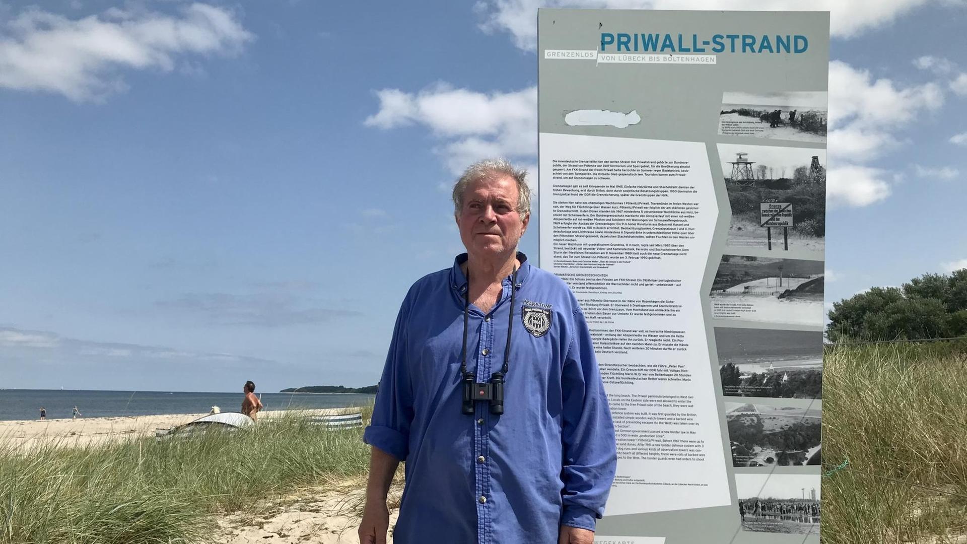 Günter Wosnitza hat graue Haare, ein blaues Hemd und steht am Strand vor einer Gedenktafel.