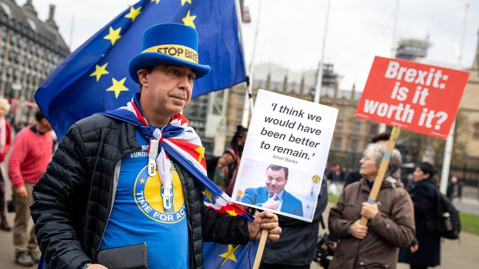 Ein Mann demonstriert mit anderen Brexit-Gegnern auf dem Londoner Parliament Square. Er trägt einen blauen Zylinder mit der Aufschrift: "Stop Brexit". Außerdem hält er ein Plakat mit einem Zitat von Arron Banks hoch: "I think we would have been better to remain." Im Hintergrund ist eine Frau zu sehen, die ein Plakat mit der Aufschrift hochhält: "Brexit: is it worth it?"
