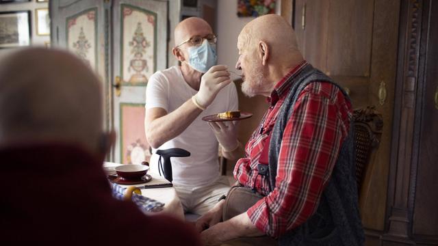 Ein Pfleger mit Mundschutz füttert einen alten Mann.