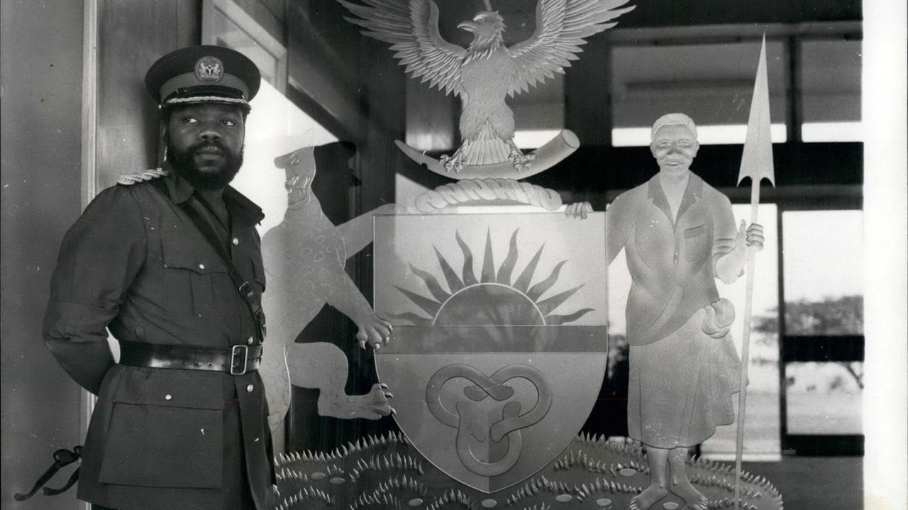 Ein historisches Schwarz-Weiß-Foto. Zu sehen ist links ein afrikanischer Mann mit Vollbart und in Militäruniform. Er steht vor dem Symbol des damaligen Staates Biafra im Südosten von Nigeria. Das Symbol zeigt unter anderem einen Adler und eine aufgehende Sonne. Ein historisches Schwarz-Weiß-Foto. Zu sehen ist links ein afrikanischer Mann mit Vollbart und in Militäruniform. Er steht vor dem Symbol des damaligen Staates Biafra im Südosten von Nigeria. Das Symbol zeigt unter anderem einen Adler und eine aufgehende Sonne.