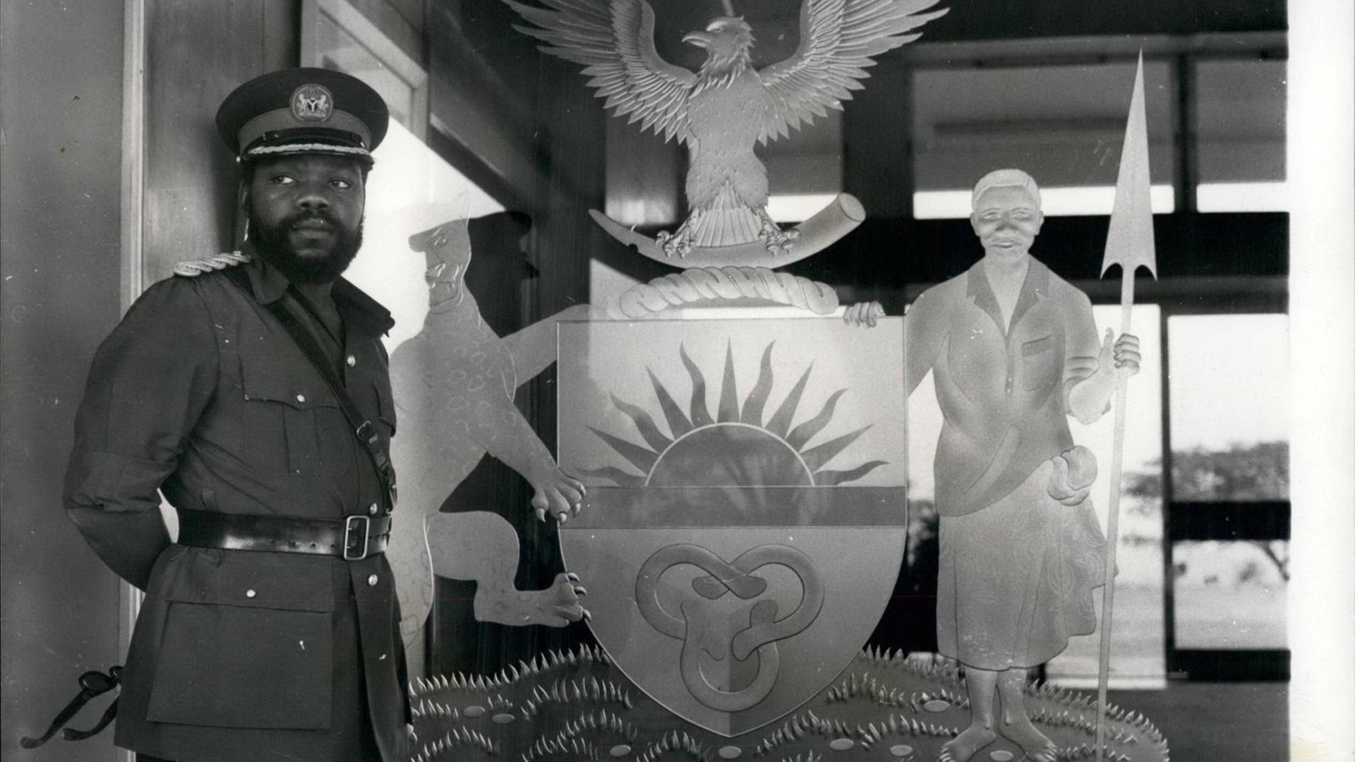 Ein historisches Schwarz-Weiß-Foto. Zu sehen ist links ein afrikanischer Mann mit Vollbart und in Militäruniform. Er steht vor dem Symbol des damaligen Staates Biafra im Südosten von Nigeria. Das Symbol zeigt unter anderem einen Adler und eine aufgehende Sonne.