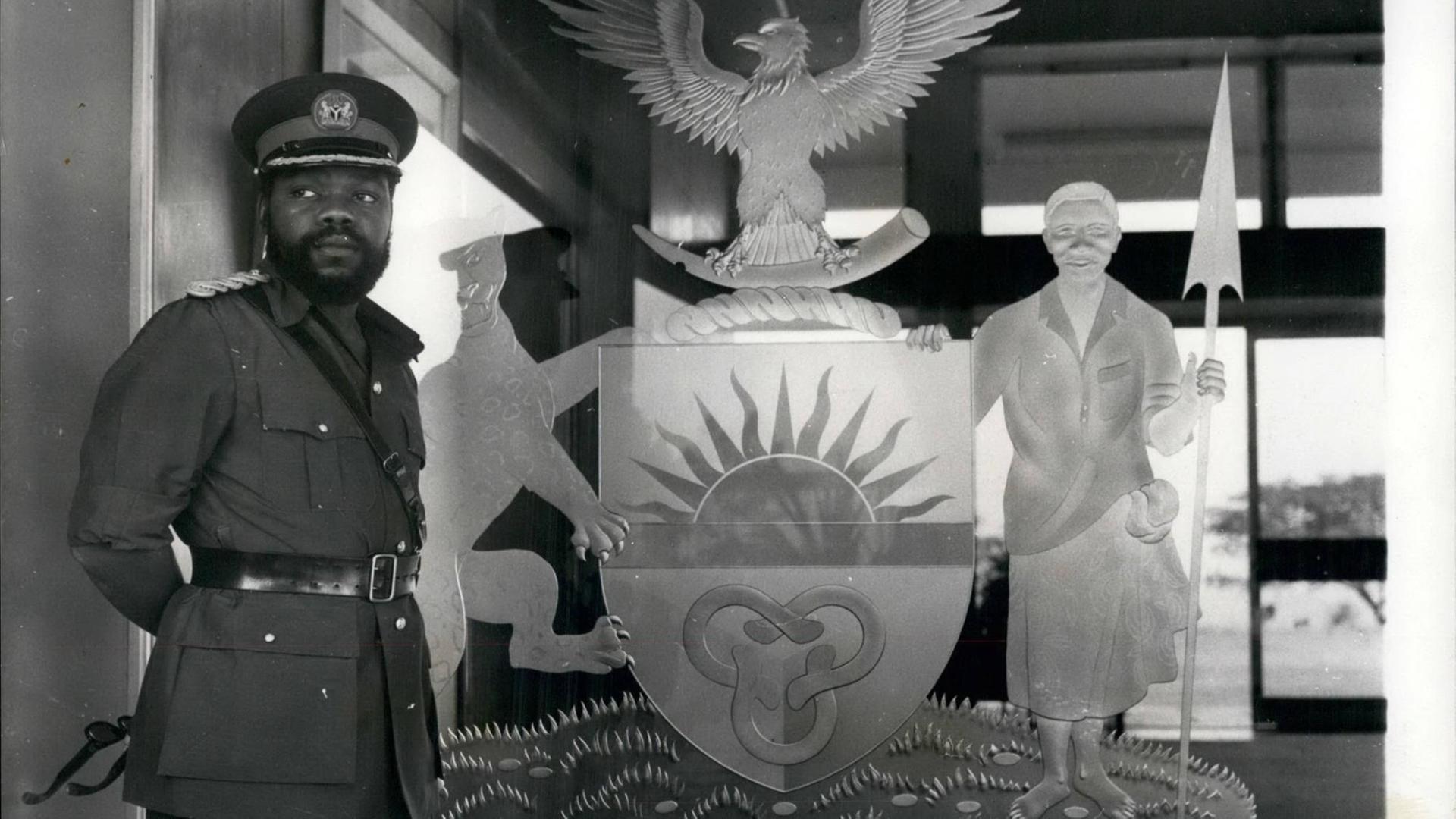 Ein historisches Schwarz-Weiß-Foto. Zu sehen ist links ein afrikanischer Mann mit Vollbart und in Militäruniform. Er steht vor dem Symbol des damaligen Staates Biafra im Südosten von Nigeria. Das Symbol zeigt unter anderem einen Adler und eine aufgehende Sonne. Ein historisches Schwarz-Weiß-Foto. Zu sehen ist links ein afrikanischer Mann mit Vollbart und in Militäruniform. Er steht vor dem Symbol des damaligen Staates Biafra im Südosten von Nigeria. Das Symbol zeigt unter anderem einen Adler und eine aufgehende Sonne.