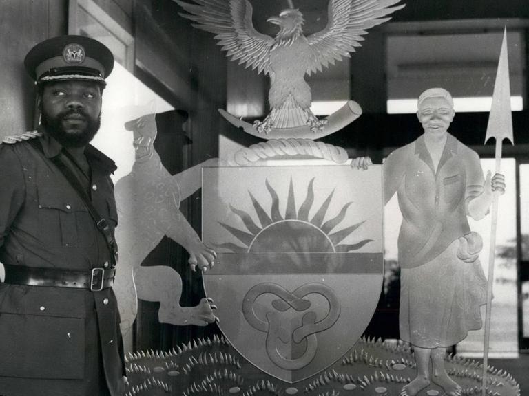 Ein historisches Schwarz-Weiß-Foto. Zu sehen ist links ein afrikanischer Mann mit Vollbart und in Militäruniform. Er steht vor dem Symbol des damaligen Staates Biafra im Südosten von Nigeria. Das Symbol zeigt unter anderem einen Adler und eine aufgehende Sonne.
