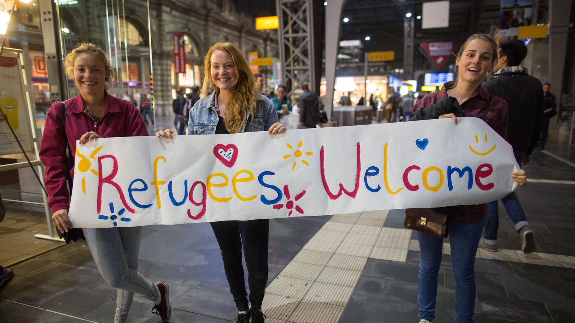 Drei junge Frauen stehen am 05.09.2015 in Frankfurt am Main (Hessen) im Hauptbahnhof mit einem Begrüßungsplakat mit der Aufschrift "Refugees Welcome" für Flüchtlinge an den Gleisen. Foto: Frank Rumpenhorst Drei junge Frauen stehen am 05.09.2015 in Frankfurt am Main (Hessen) im Hauptbahnhof mit einem Begrüßungsplakat mit der Aufschrift "Refugees Welcome" für Flüchtlinge an den Gleisen. Foto: Frank Rumpenhorst