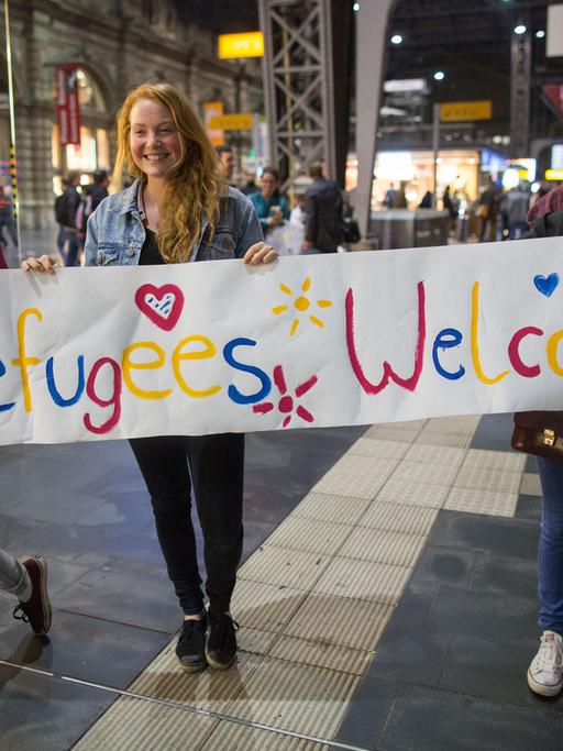 Drei junge Frauen stehen am 05.09.2015 in Frankfurt am Main (Hessen) im Hauptbahnhof mit einem Begrüßungsplakat mit der Aufschrift "Refugees Welcome" für Flüchtlinge an den Gleisen. Foto: Frank Rumpenhorst