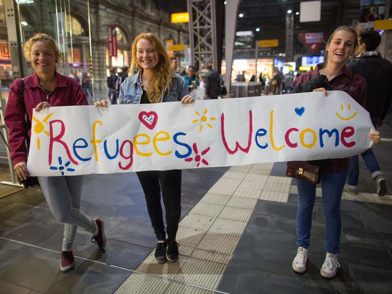 Drei junge Frauen stehen am 05.09.2015 in Frankfurt am Main (Hessen) im Hauptbahnhof mit einem Begrüßungsplakat mit der Aufschrift "Refugees Welcome" für Flüchtlinge an den Gleisen. Foto: Frank Rumpenhorst