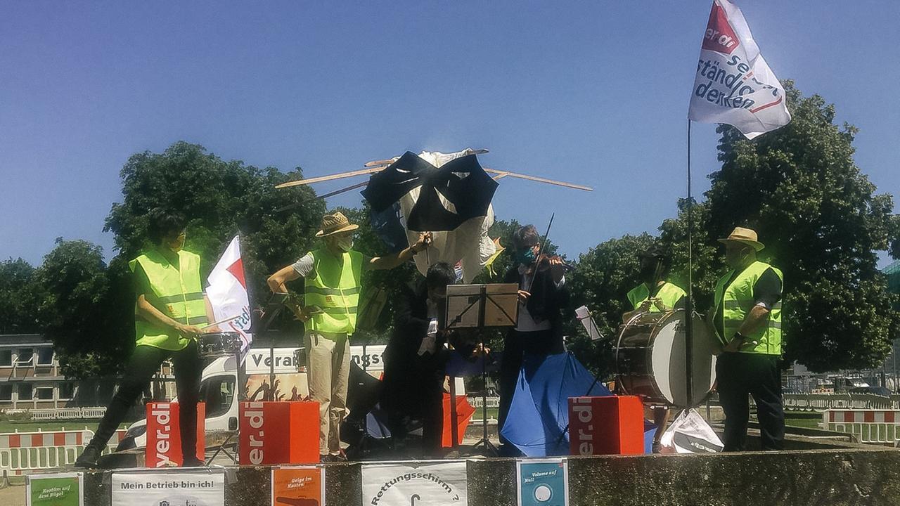 Musiker in Warnwesten und mit Mundschutz demonstrieren mit löchrigen Schirmen und Musik auf einer Bühne im Freien. Musiker in Warnwesten und mit Mundschutz demonstrieren mit löchrigen Schirmen und Musik auf einer Bühne im Freien.